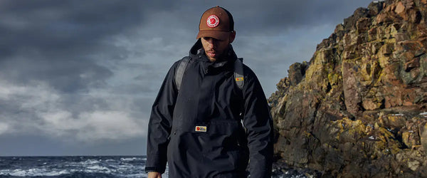 Man wearing a Fjällräven jacket and beanie walking along a rocky shoreline with the ocean in the background.