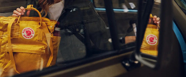A person in a car holding a yellow Fjallraven Kanken backpack with a visible red fox logo, reflected in the vehicle’s door pocket.