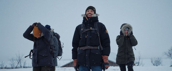 Group of friends trekking in a snowy landscape, wearing a Fjällräven expedition down jacket with prominent branding, highlighting the warmth and quality of the outerwear.