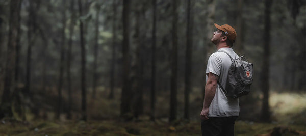 Photographer Brad Thomas, sporting a cap and a grey Fjällräven Kånken backpack, looks up thoughtfully into the forest canopy.