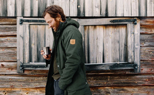 Man leaning against a rustic wooden wall, enjoying a warm drink and wearing a Fjällräven Greenland jacket in deep green.