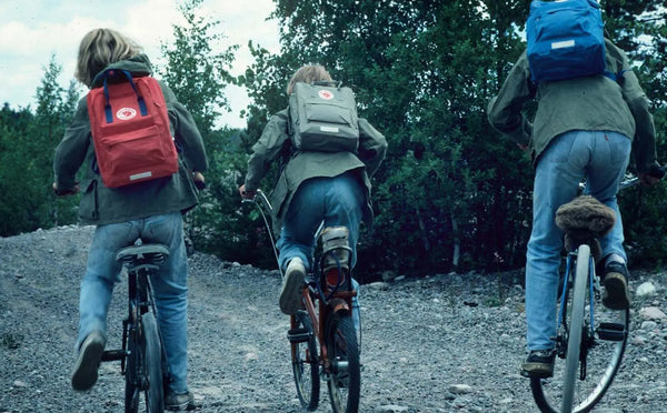 Three cyclists riding on a rough trail with Fjällräven backpacks in red, grey, and blue
