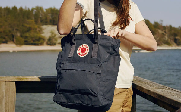 Close-up of a woman holding a Fjällräven Kånken tote bag in navy blue with the classic red and white logo, against a coastal backdrop.