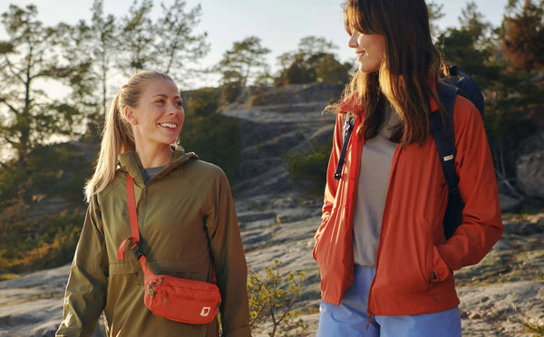 Friends enjoying an outdoor hike, one wearing a Fjällräven green jacket with a matching hip pack and the other in an orange Fjällräven jacket with a navy backpack.