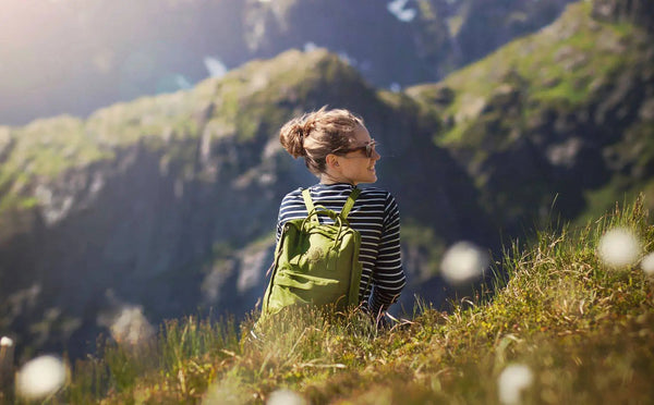 Woman seated in mountain grass with a Fjällräven Kånken backpack in vibrant green.
