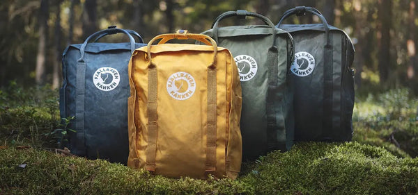 Four Fjällräven Tree-Kånken backpacks in blue, yellow, and two shades of green, displayed in a forest setting on moss-covered ground.