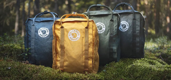 Four Fjällräven Kånken backpacks in blue, yellow, and two shades of green, displayed in a forest setting on moss-covered ground.