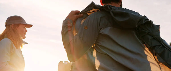 Back view of an adventurer wearing a Fjällräven jacket with a logo visible on the sleeve, assisting a fellow hiker adjusting a cap against a warm sunset backdrop.
