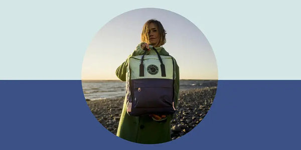 Woman in green coat holding a purple and green Kanken backpack on a pebble beach at sunset.