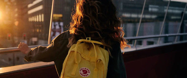 Woman with curly hair wearing a Fjällräven yellow backpack looking over the city from a bridge at sunset.