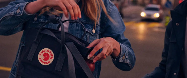 Woman opening a Fjällräven Kånken sling bag to retrieve an item, with the distinctive Fjällräven logo visible, on a busy street during twilight.