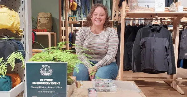 Smiling woman sitting at a Fjällräven store during a custom embroidery event, with a sign and embroidery materials on the table.